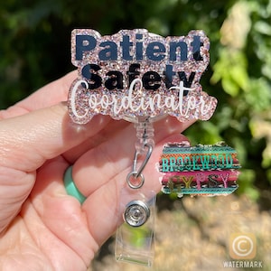 May include: A pink glitter badge holder with the text "Patient Safety Coordinator" in black lettering. The badge holder is attached to a clear retractable badge reel with a silver clip. A smaller, colorful patterned badge holder with the text "Brookwood Gypsy" is attached to the reel.