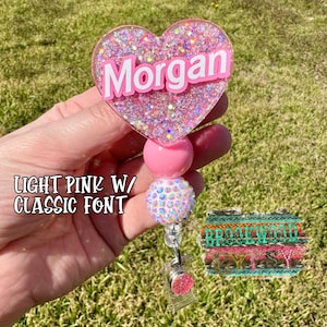 May include: A light pink heart-shaped badge holder with the name "Morgan" in white letters. The holder is decorated with glitter and has a pink and white beaded chain.