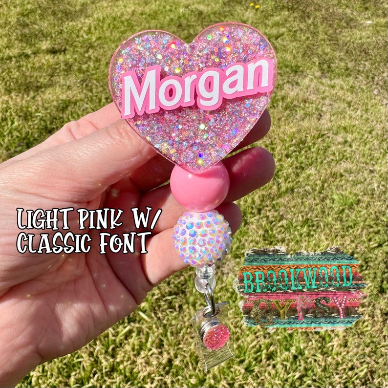 May include: A light pink heart-shaped badge holder with the name "Morgan" in white letters. The holder is decorated with glitter and has a pink and white beaded chain.