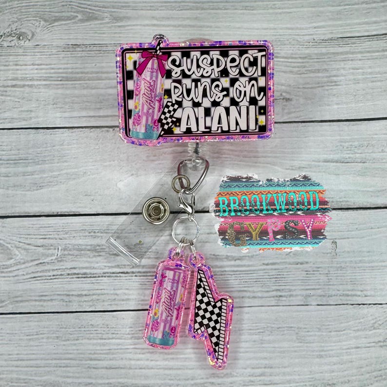 May include: A retractable badge reel with the text "suspect puns on Alani". The reel has a pink and black checkered flag design and a pink bow. A smaller charm with a pink and black checkered flag design is attached.
