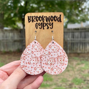May include: A pair of teardrop-shaped earrings with a white background and a red ghost pattern. The earrings are made of cork and have silver hooks. The earrings are displayed on a wooden stand with the text "Brookwood Gypsy" engraved on it.