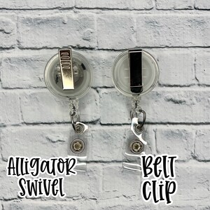 Puede incluir: Dos portagajes retr&aacute;ctiles de pl&aacute;stico transparente con clips met&aacute;licos. El portagajes de la izquierda tiene un clip giratorio de cocodrilo y el portagajes de la derecha tiene un clip de cintur&oacute;n. Ambos portagajes est&aacute;n colgados de una pared de ladrillo blanco.