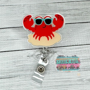 May include: A red crab wearing blue sunglasses and sitting on a tan beach is attached to a clear retractable badge holder. The badge holder has a white stripe pattern and a silver button.