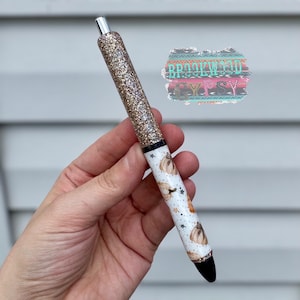 May include: A glittery gold pen with a black tip and silver accents. The pen's barrel features a white background with a pattern of pumpkins and stars. The pen has the text "BROOKWOOD" on it.