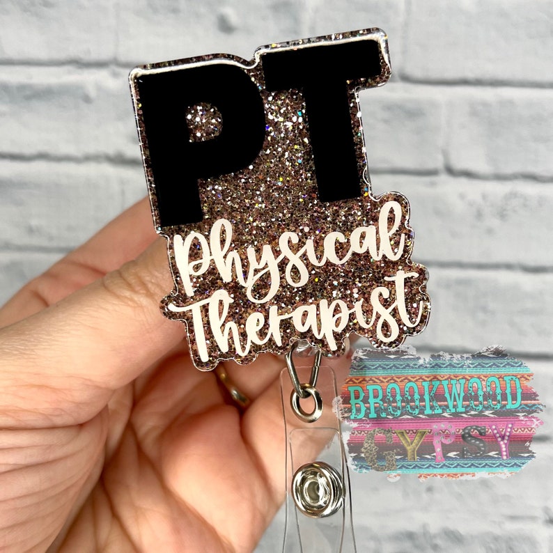 May include: A gold glitter badge holder with a black "PT" and the text "Physical Therapist" in white. The badge holder is attached to a clear retractable badge reel.