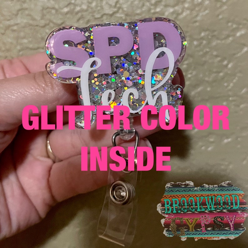 Puede incluir: Un portagafete de purpurina morado y blanco con el texto "SPD Tech" en el interior. El portagafete tiene un clip de pl&aacute;stico transparente en la parte posterior.