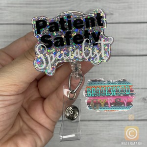 May include: A silver glitter badge holder with a black text overlay that reads "Patient Safety Specialist". The badge holder is attached to a clear retractable badge reel with a silver clip. A colorful patterned fabric with the text "Brookwood Gypsy" is visible in the background.