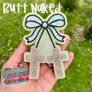 May include: A light green and glittery cross-shaped accessory with a bow and the text "Butt Naked". The accessory has two glittery crosses hanging from the bow. The bow is light blue with black outlines.