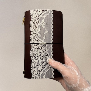May include: A dark brown leather journal cover featuring a wide vertical strip of white floral lace down the center. A thin black elastic band secures the lace, accented with three small white faux pearls. A gold metal charm hangs from the side.