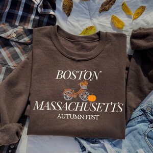 May include: Brown sweatshirt with white text that reads "Boston, Massachusetts' Autumn Fest". The sweatshirt features a graphic of a bicycle with a basket of flowers and a pumpkin.