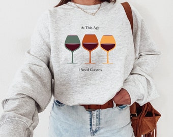 May include: A gray sweatshirt with the text "At This Age I Need Glasses" and a graphic of three wine glasses with different colored liquid.