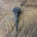May include: A hand-forged iron nail with a square head and a pointed tip. The nail is resting on a piece of wood.