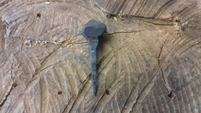 May include: A hand-forged iron nail with a square head and a pointed tip. The nail is resting on a piece of wood.