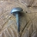 May include: A single, hand-forged nail with a rounded, domed head. The nail is made of metal and has a dark gray finish.