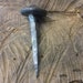 May include: A hand-forged nail with a large, flat head. The nail is made of metal and has a rustic, aged appearance. It is sitting on a piece of wood.