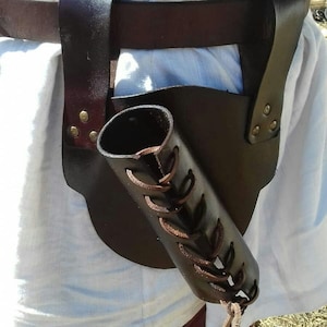 Puede incluir: Un carcaj de cuero marrón oscuro con un diseño cilíndrico, sujeto a un cinturón y una correa para el hombro. El carcaj presenta cordones decorativos y está asegurado con remaches. El diseño sugiere un tema medieval o histórico.