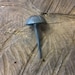 May include: A single, dark gray, metal nail with a rounded, domed head. The nail is sitting on a piece of rough, brown wood.
