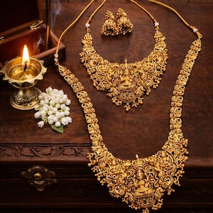 Op de afbeelding: Een sierlijke, goudkleurige ketting- en oorbellenset met ingewikkelde details. De sieraden hebben een centraal figuur en uitgebreide patronen. De set wordt getoond op een donkerhouten oppervlak, samen met een brandende kaars en bloemen.