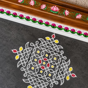 May include: A dark gray floor surface features a white, yellow, and red geometric kolam pattern. Above the pattern, a wooden border is painted with a row of pink lotus flowers and green vines, followed by a white border with pink lotus buds.