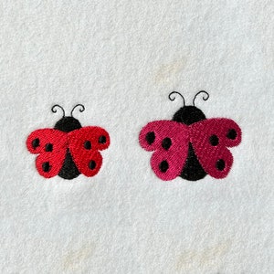 Könnte beinhalten: Zwei gestickte Marienkäfer, einer rot und einer rosa, mit schwarzen Punkten und Fühler. Die Marienkäfer befinden sich auf einem weißen Hintergrund.