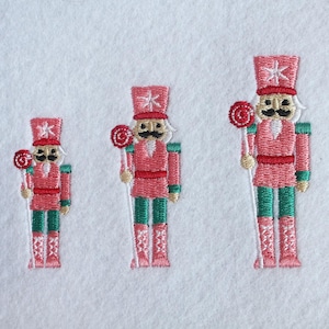 May include: Three embroidered nutcracker figures of varying sizes arranged in a row on a white fabric background. Each nutcracker wears a pink uniform with green trousers, a tall pink hat with a white star, and holds a red and white lollipop.