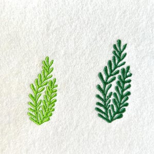 May include: Two embroidered green sprigs on a white background. The sprig on the right is larger and darker green than the sprig on the left.