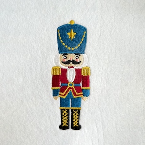 May include: Embroidered patch of a toy soldier nutcracker. The nutcracker has a blue hat with a gold star, a white face, a black mustache, and a red and blue uniform with gold accents. The background is white.