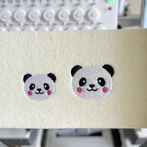 Puede incluir: Dos parches bordados de osos panda. Los pandas tienen caras blancas, orejas y ojos negros y mejillas rosas. Los parches están sobre una tela color crema, probablemente para coser o hacer manualidades. El fondo incluye una máquina de bordar.