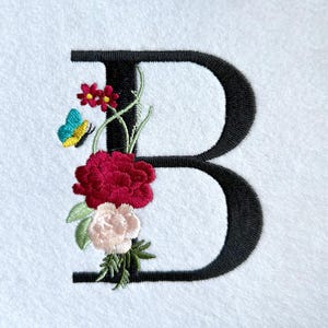 May include: Embroidered black letter "B" adorned with a floral design. The flowers include a large red peony, a light pink bloom, and small red flowers. A small butterfly with blue and yellow wings adds a whimsical touch.