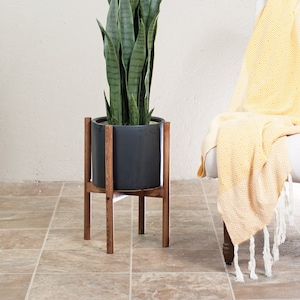 May include: A potted snake plant on a wooden plant stand. The plant is in a dark gray pot and the stand is made of dark brown wood. The plant is in a room with a white wall and a yellow blanket on a chair in the background.