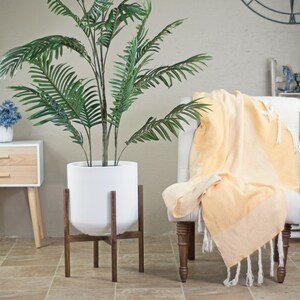 May include: A white planter with a wooden stand, holding a large green artificial palm tree. The planter is sitting on a tiled floor in a room with a white chair and a yellow throw blanket.