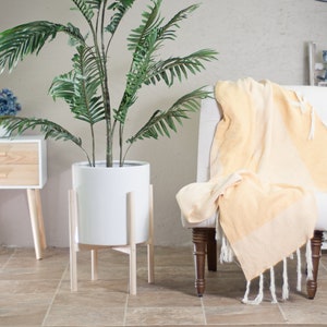 May include: A white ceramic planter with a wooden stand, holding a green potted plant. The plant is a large, leafy palm tree. The planter is sitting on a tiled floor in front of a white wall. A yellow throw blanket is draped over a chair with wooden legs.