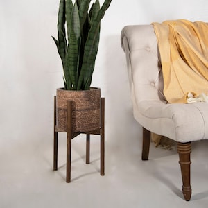 May include: A potted snake plant with long, green leaves sits on a wooden plant stand. The plant is in a brown pot with a textured surface. A white chair with a yellow throw blanket is in the background.