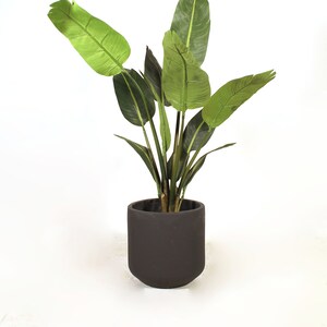 May include: A green artificial bird of paradise plant in a dark gray pot. The plant has multiple leaves and is on a white background.