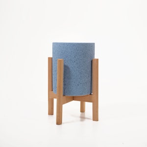 May include: A light blue ceramic planter with a speckled finish sits on a wooden stand with four legs. The stand is made of natural wood and has a simple, rustic design.