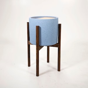 May include: A light blue speckled ceramic planter pot on a dark brown wooden plant stand. The cylindrical pot has a wide opening and sits securely within the stand's frame. The stand has four legs and a cross-beam support.