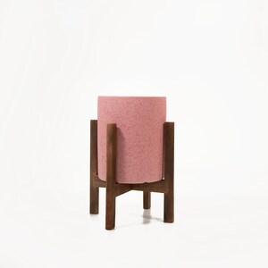 May include: A pink ceramic planter with a textured surface sits on a wooden stand with four legs. The stand is made of dark brown wood.