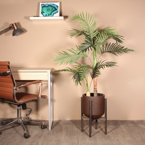 May include: A brown ceramic planter with a black metal stand, holding a green artificial palm tree. The planter is on a wooden floor in front of a desk with a brown leather office chair.