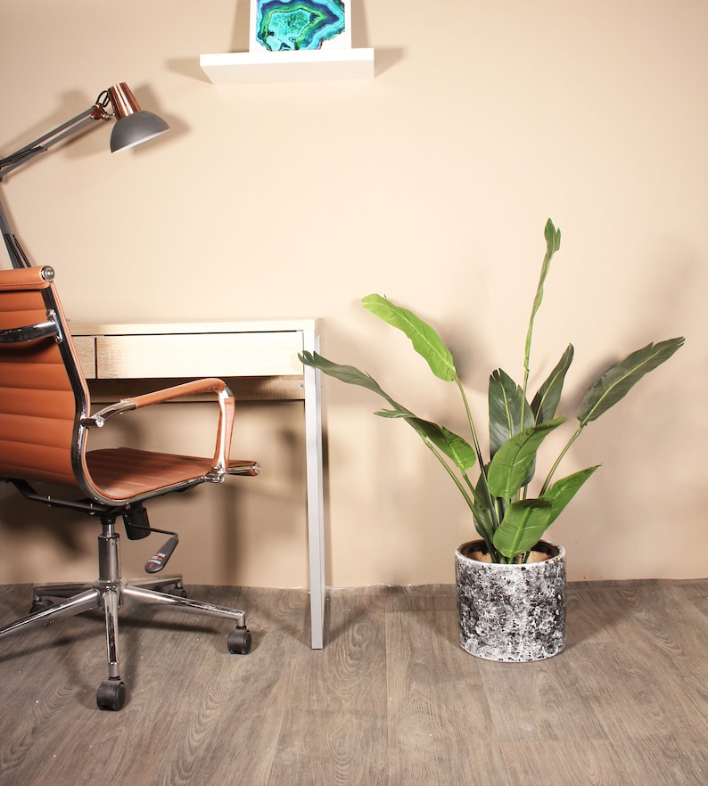 May include: A brown leather office chair with chrome arms and base sits in front of a white desk with a potted green plant to the right. The plant is in a white and gray speckled pot. A desk lamp with a copper base and a white shade is on the wall behind the chair.
