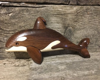 May include: A handcrafted wooden orca sculpture. The orca is made of dark brown wood with white accents and a smooth finish. The sculpture is lying on a weathered wooden surface. The orca's fins and tail are detailed.