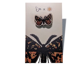 May include: A wooden butterfly pin with a black and orange pattern, displayed on a card with a matching butterfly illustration. The card has the text "Ejs x" and a sun graphic.