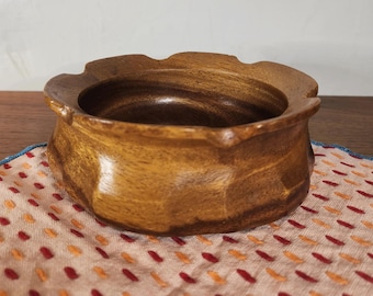 Könnte beinhalten: Eine runde, mittelbraune Holzschale mit glatter, polierter Oberfläche und einem gewellten, geschwungenen Rand. Die Außenseite weist ein facettiertes, mehrseitiges Design auf, das für ein strukturiertes Aussehen sorgt.
