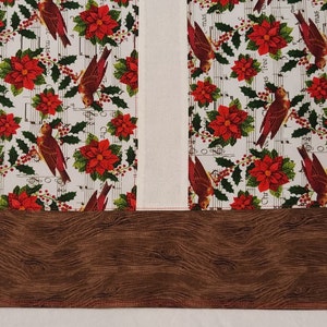 May include: A white fabric with a repeating pattern of red poinsettias, green holly leaves, and brown birds singing Christmas carols. The fabric has a brown woodgrain border.