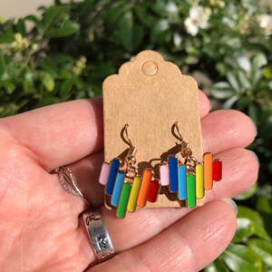 May include: Gold-tone earrings with colorful, vertical bar designs in shades of pink, blue, green, yellow, orange, and red. The earrings are displayed on a brown card, with a green leafy background.
