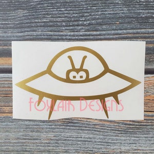 May include: Gold vinyl sticker of a cartoon UFO with a simple face. The sticker is on a white background.