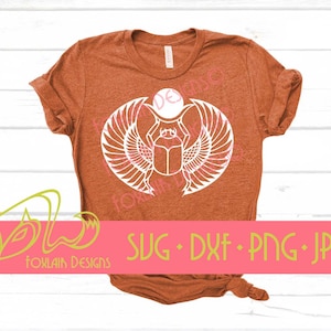 May include: An orange t-shirt featuring a white graphic of a scarab beetle with wings, a sun disc, and the text "SVG • DXF • PNG • JPG". The shirt has a crew neck and short sleeves. The design is centered on the chest.