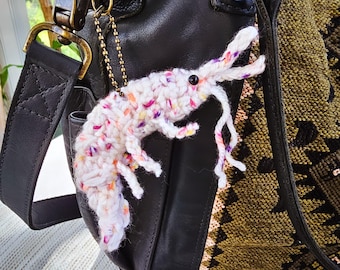 May include: A white crocheted shrimp keychain with pink, purple, and orange accents hanging from a black leather bag with a brown strap.