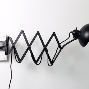 May include: A black, articulated wall-mounted lamp with a round shade. The lamp has a scissor-like arm that extends from a wall mount. The shade is a semi-circle shape. The lamp is designed for directional lighting.