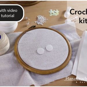 May include: Crochet kit with a white embroidery hoop, white fabric, white yarn, and white felt circles. The kit includes a video tutorial. "Crochet kit" and "Hana Crochet & design" are written on the image.