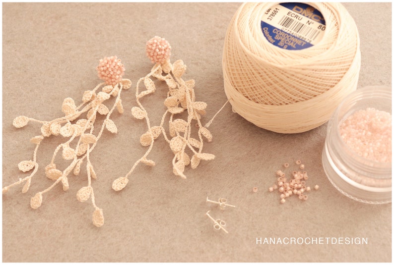 May include: A pair of delicate crocheted earrings with pink bead accents. The earrings are made with a beige crochet thread and feature a cascading design. The image also shows a spool of beige crochet thread, a small container of pink beads, and two earring backs.